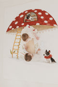 Two children playing on a large red and white polka dot umbrella with a ladder and small house design.